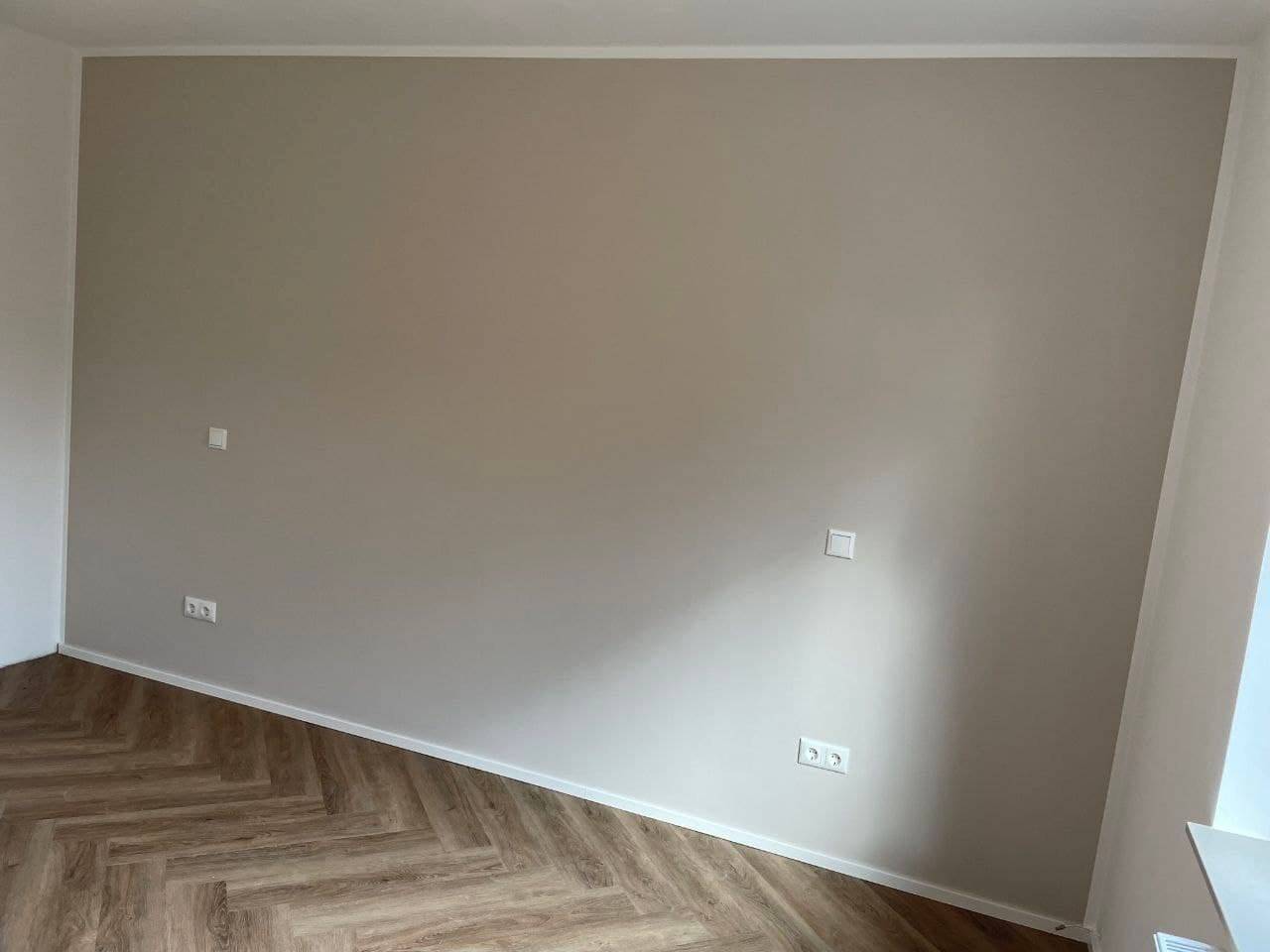 Große Wandfläche in warmem Greige, sauber gegen weiße Decke und Nachbarwand abgesetzt, davor ein Designboden im Fischgrätmuster