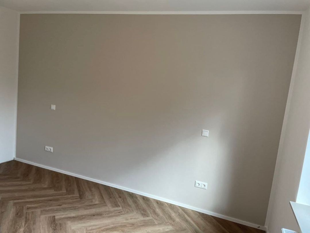 Große Wandfläche in warmem Greige, sauber gegen weiße Decke und Nachbarwand abgesetzt, davor ein Designboden im Fischgrätmuster